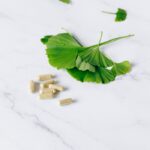 Flat lay of herbal capsules and fresh ginkgo leaves on a marble surface for natural remedy concept.